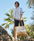 A person stands outdoors on rocks holding a beige and white Westford Mill WM451 tote bag, wearing a white shirt and blue shorts, with a palm tree and clear blue sky in the background.