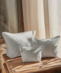 Three Westford Mill WM540 white canvas zipper pouches of varying sizes are placed on a wooden surface in front of a window with sheer curtains.