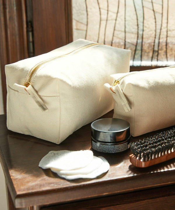 Two beige WM552 toiletry bags from Westford Mill with zippers are placed on a wooden table beside a small tin, a hairbrush, and round cotton pads.