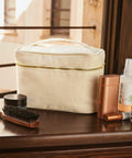 A beige Westford Mill WM558 toiletry bag sits on a wooden surface with a hairbrush, compact jar, brown cylindrical container, copper-colored rectangular bottle, and a spray bottle next to it.