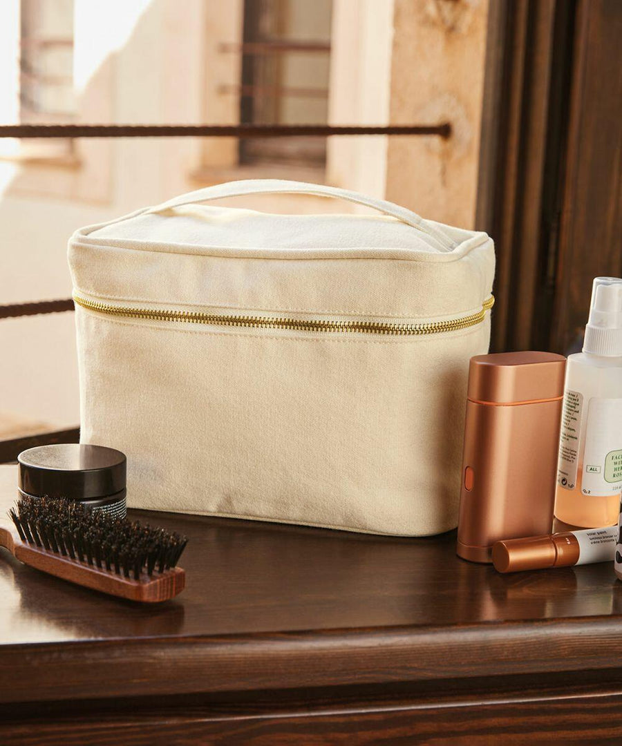 A beige Westford Mill WM558 toiletry bag sits on a wooden surface with a hairbrush, compact jar, brown cylindrical container, copper-colored rectangular bottle, and a spray bottle next to it.