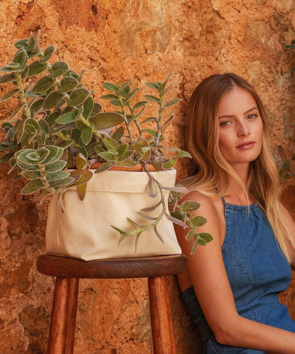 A woman in a blue denim dress sits against a textured wall. Beside her, a fabric pot from Westford Mill containing a lush, green plant rests on a wooden stool.