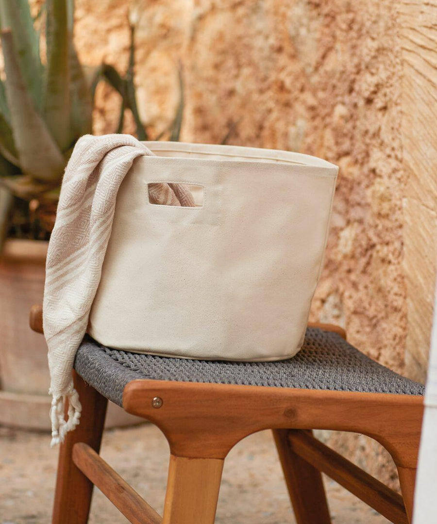 A white Westford Mill WM574 basket with a handle sits on a woven chair beside a plant in a pot, placed against a textured beige wall. A light beige towel is partially draped over the basket's edge.