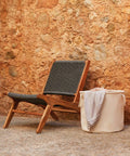 A wicker chair with wooden frame is placed against a textured stone wall. A cream-colored Westford Mill WM580 fabric storage basket containing folded textiles sits next to the chair.