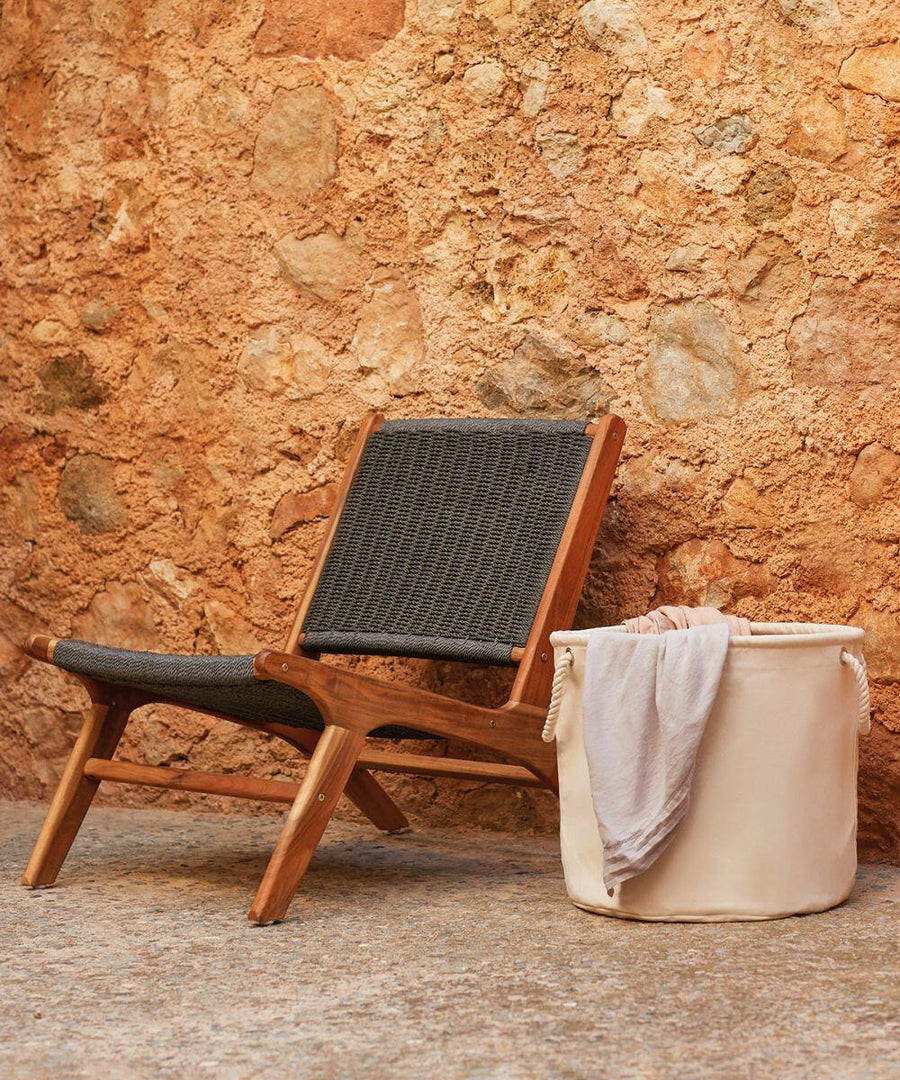 A wicker chair with wooden frame is placed against a textured stone wall. A cream-colored Westford Mill WM580 fabric storage basket containing folded textiles sits next to the chair.