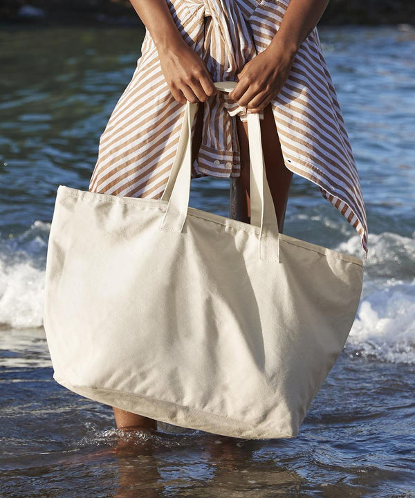 Standing in shallow beach water, a person holds a Westford Mill oversized heavy duty canvas tote bag. They wear a striped wrap skirt, with small waves visible in the background.