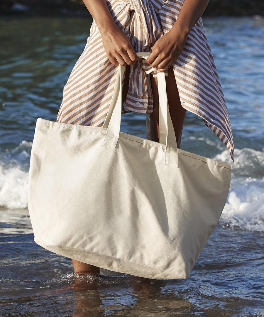 Standing in shallow beach water, a person holds a Westford Mill oversized heavy duty canvas tote bag. They wear a striped wrap skirt, with small waves visible in the background.