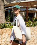 A person with a green cap and white shirt carries a beige Westford Mill WM610 while walking outdoors near umbrellas and plants.
