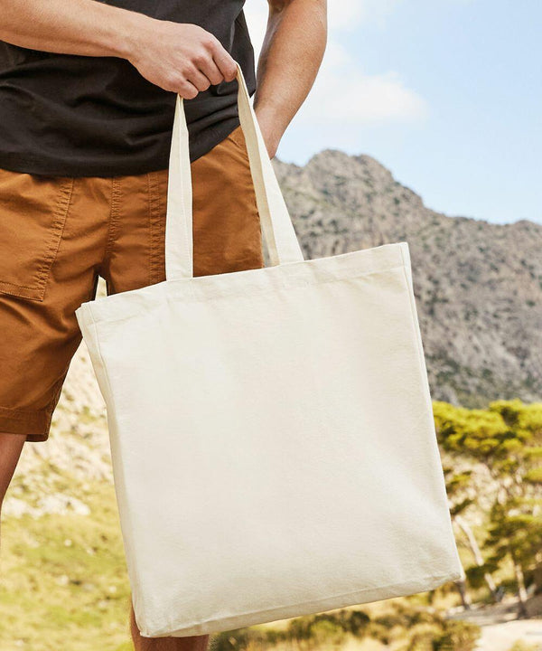 Person in an outdoor setting holding a large beige WM623 tote bag by Westford Mill by its handles, wearing a black shirt and brown shorts. Mountainous landscape is visible in the background.