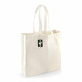 A plain white canvas tote bag from Westford Mill, identified as product WM623, stands upright with two handles. Attached to the top of the bag near the handles is a small black tag featuring green and white text.