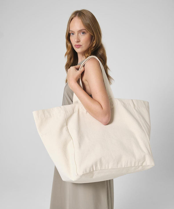 A woman with long, light brown hair holds the Westford Mill Oversized beach bag over her shoulder. She wears a sleeveless, light-coloured dress and stands against a plain grey background.