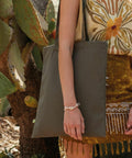 Person holding a large olive green WM801 tote bag from Westford Mill, standing next to a cactus, wearing a patterned shirt and a bracelet.