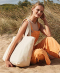 A woman in orange overalls and beige top sits on sandy ground holding a large Westford Mill WM855 tote bag, with tall grass in the background.
