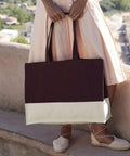 A person in a light pink dress stands outdoors on a stone ledge, holding the Westford Mill EarthAware organic contrast shopper tote in dark brown and white, and wearing light-coloured espadrille shoes.