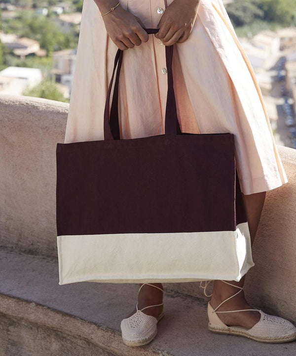 A person in a light pink dress stands outdoors on a stone ledge, holding the Westford Mill EarthAware organic contrast shopper tote in dark brown and white, and wearing light-coloured espadrille shoes.