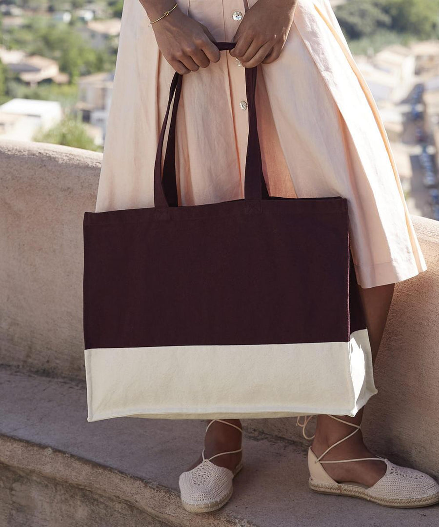 A person in a light pink dress stands outdoors on a stone ledge, holding the Westford Mill EarthAware organic contrast shopper tote in dark brown and white, and wearing light-coloured espadrille shoes.