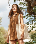 A woman with curly hair, wearing a brown sleeveless dress and carrying a beige Westford Mill WM901 tote bag, stands outdoors next to a tree.