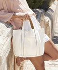 A person outdoors leans against a stone wall, wearing a light pink long-sleeve blouse and white shorts whilst holding the Westford Mill Courtside medium tote, a white canvas bag with brown trim.