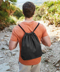 A person in an orange shirt and light-colored pants is seen from behind, walking on a rocky path while wearing a black Westford Mill WM960 drawstring backpack. Trees and foliage are visible in the background.