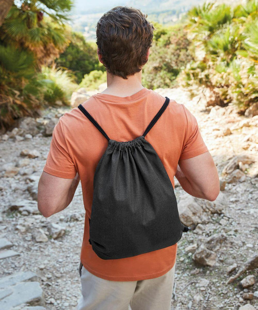 A person in an orange shirt and light-colored pants is seen from behind, walking on a rocky path while wearing a black Westford Mill WM960 drawstring backpack. Trees and foliage are visible in the background.