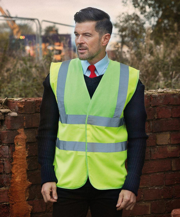 A man wearing a bright yellow Yoko YK001 safety vest over a blue shirt and red tie stands in front of a brick wall at a construction site.