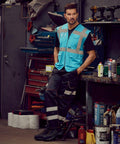 A man wearing a blue Yoko YK002 safety vest and black reflective pants stands in a workshop, leaning against a workbench with tools and equipment in the background.