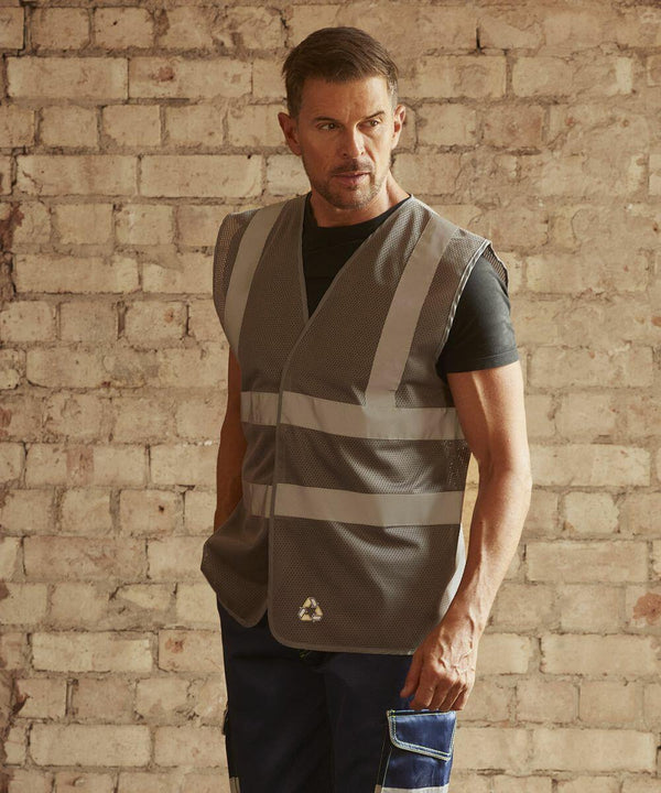 A man wearing a Yoko YK006 reflective safety vest and blue work pants with white accents stands in front of a brick wall.
