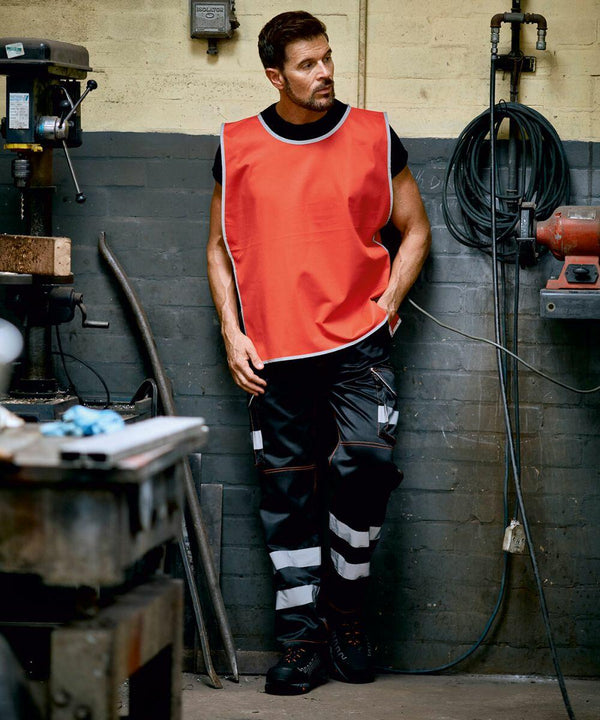 A man wearing a red Yoko YK009 safety vest and black work pants with reflective stripes stands in a workshop with various tools and machinery around him.