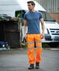 A man in a blue t-shirt and bright orange reflective YK013 Yoko pants stands outdoors near a white vehicle and a wooden structure.