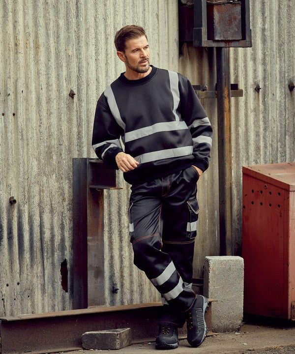 A person in Yoko YK030 high-visibility workwear leans against a corrugated metal wall in an industrial setting.