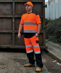 A person wearing orange and reflective safety workwear, including an orange Yoko YK034 cap, stands on a concrete surface in front of a large, rusty metal container.