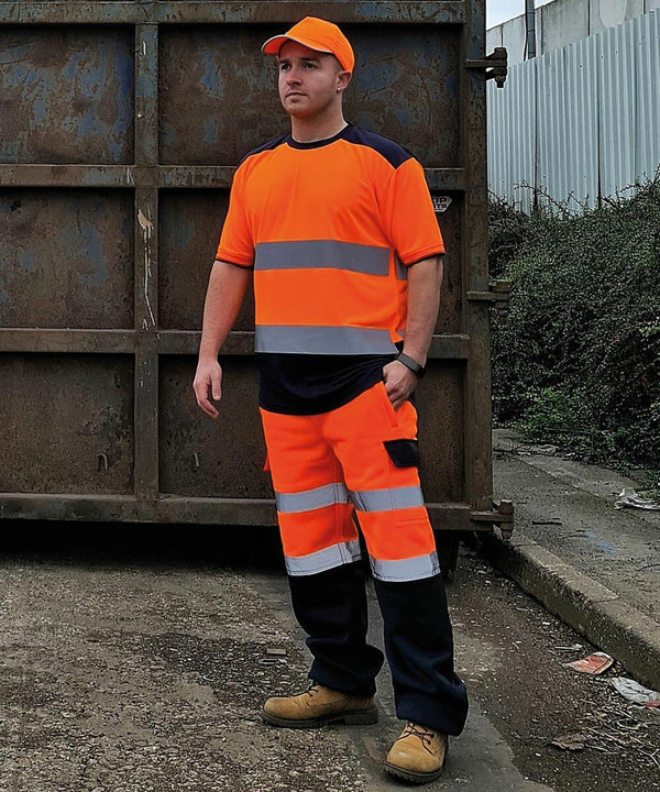 A person wearing orange and reflective safety workwear, including an orange Yoko YK034 cap, stands on a concrete surface in front of a large, rusty metal container.