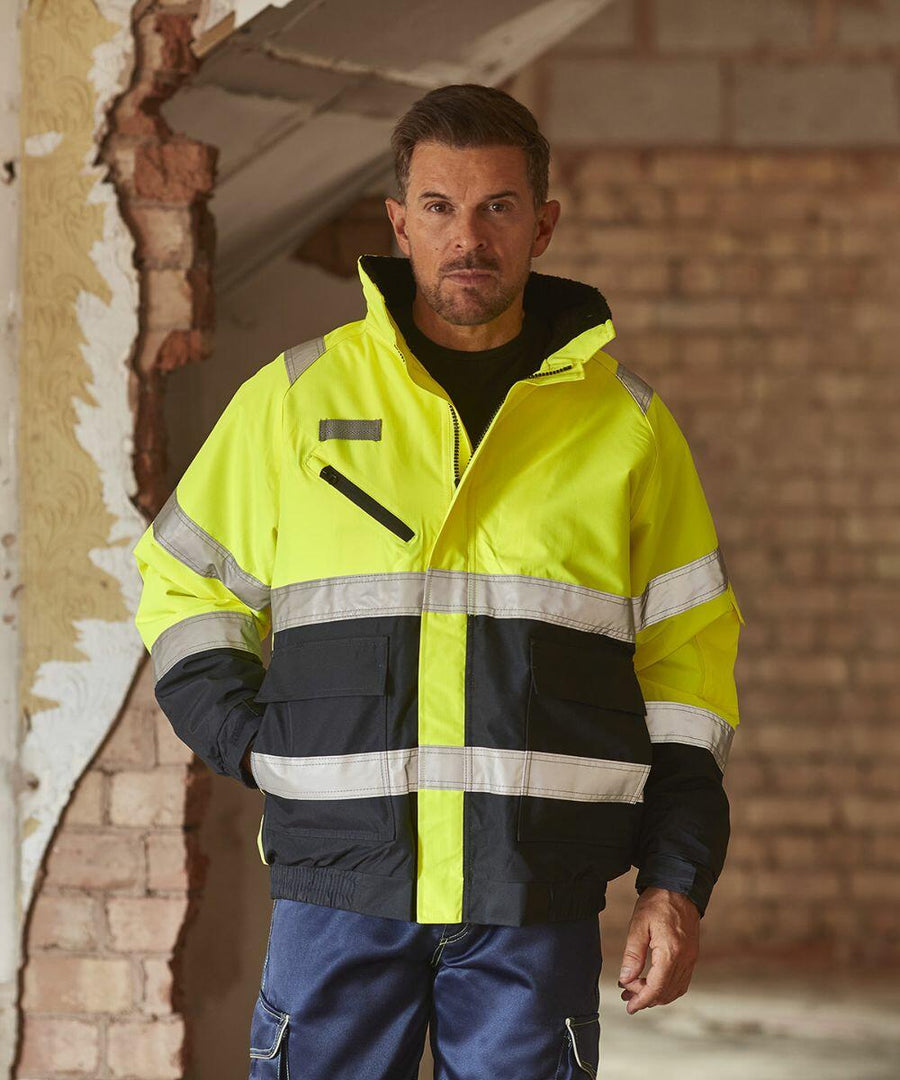 A person stands indoors wearing a high-visibility yellow and black Yoko YK035 jacket and blue pants, with hands in jacket pockets. The background shows a brick wall and a partially exposed wooden frame.