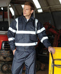 A man in a dark blue reflective work jacket and pants stands indoors, resting his hand on a yellow Yoko YK043. Heavy equipment is visible in the background.