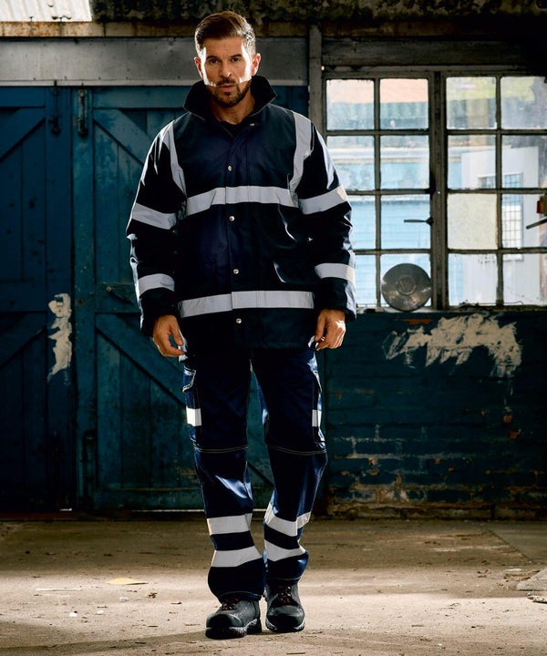 A man stands in front of a weathered building interior, wearing a Yoko YK046 safety jacket and pants with reflective stripes.