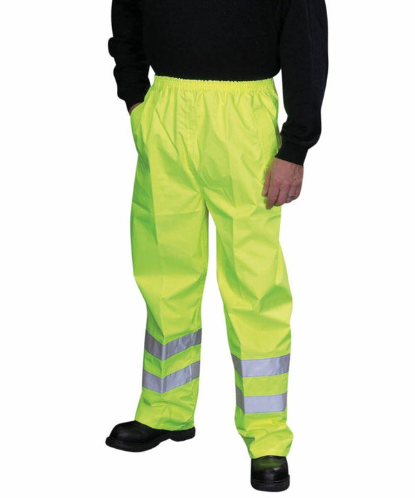 A person wearing bright yellow high-visibility Yoko YK070 pants with reflective stripes stands with hands in pockets.