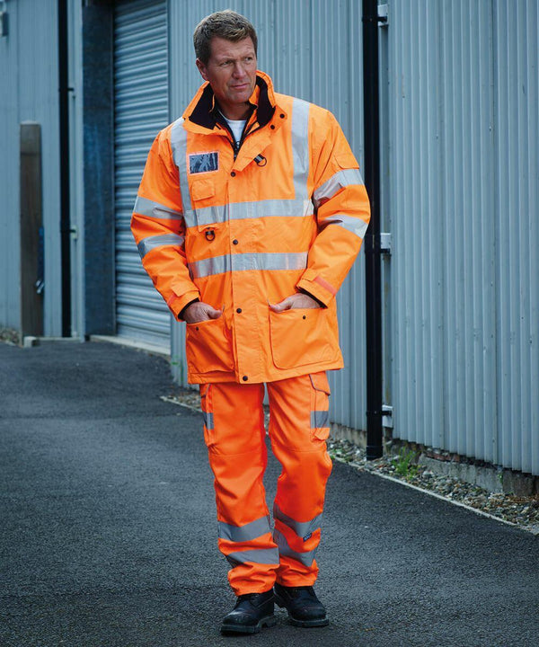 A person in a fluorescent orange Yoko YK073 jacket and pants stands outdoors near a corrugated metal wall.