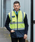A man with short hair wears a Yoko YK105 high-visibility vest over a dark sweater and collared shirt, standing in an industrial area with a shutter door in the background.