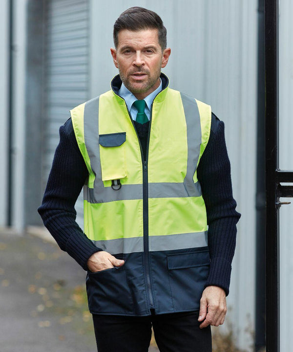 A man with short hair wears a Yoko YK105 high-visibility vest over a dark sweater and collared shirt, standing in an industrial area with a shutter door in the background.