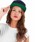 A person with long hair wears a dark Flexfit By Yupoong YP001 and a white shirt, gazing directly at the camera while holding the cap's brim with one hand.