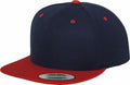 Introducing the Flexfit By Yupoong YP002: a stylish snapback cap in dark blue, featuring a vibrant red brim and accentuating red button. Designed with small red ventilation holes for breathability, it also boasts a sleek silver sticker on the brim.