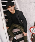 A young person wearing a black YP004 cap by Flexfit By Yupoong, a black jacket, and camouflage pants sits on outdoor stairs next to a white brick wall.