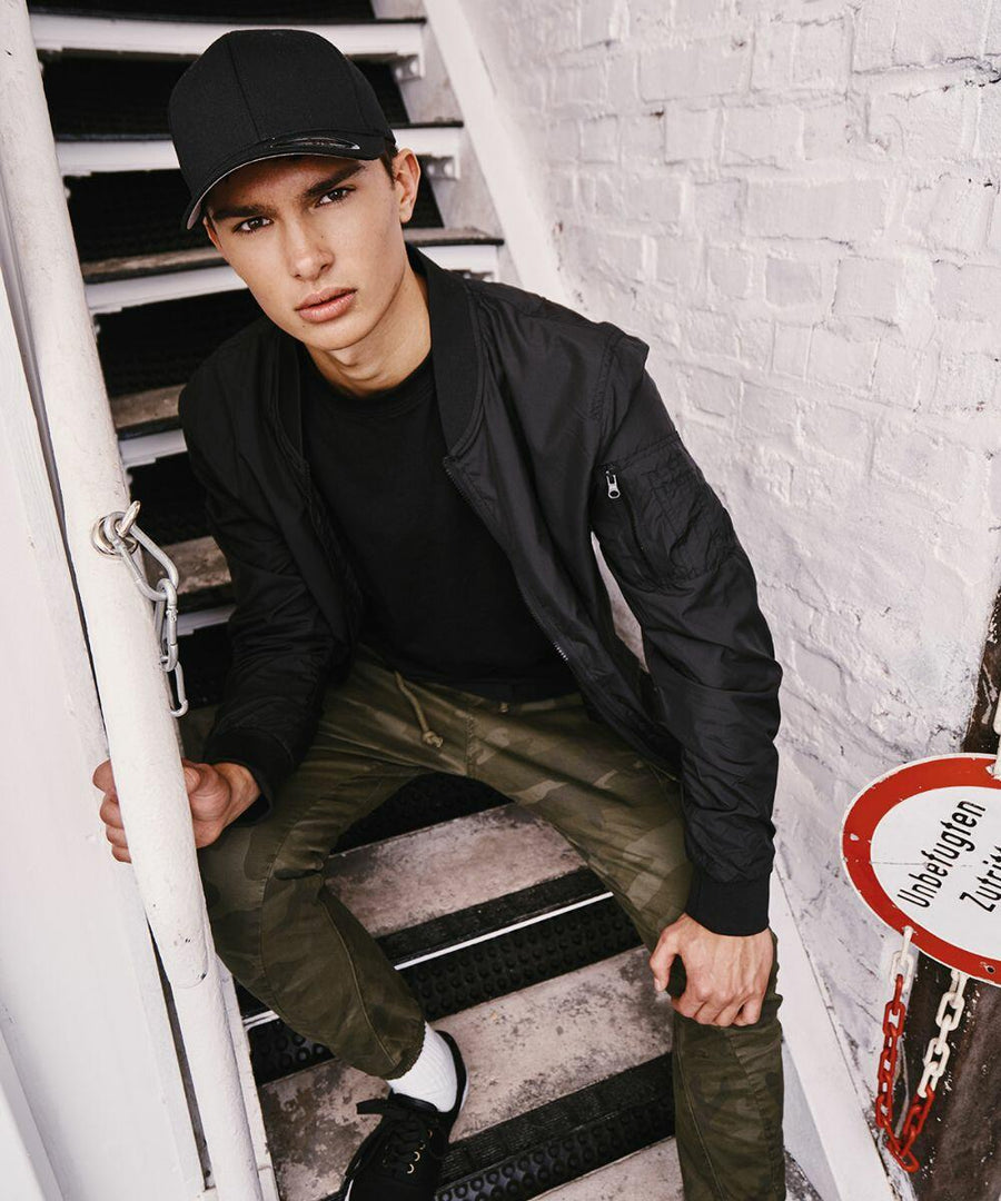 A young person wearing a black YP004 cap by Flexfit By Yupoong, a black jacket, and camouflage pants sits on outdoor stairs next to a white brick wall.