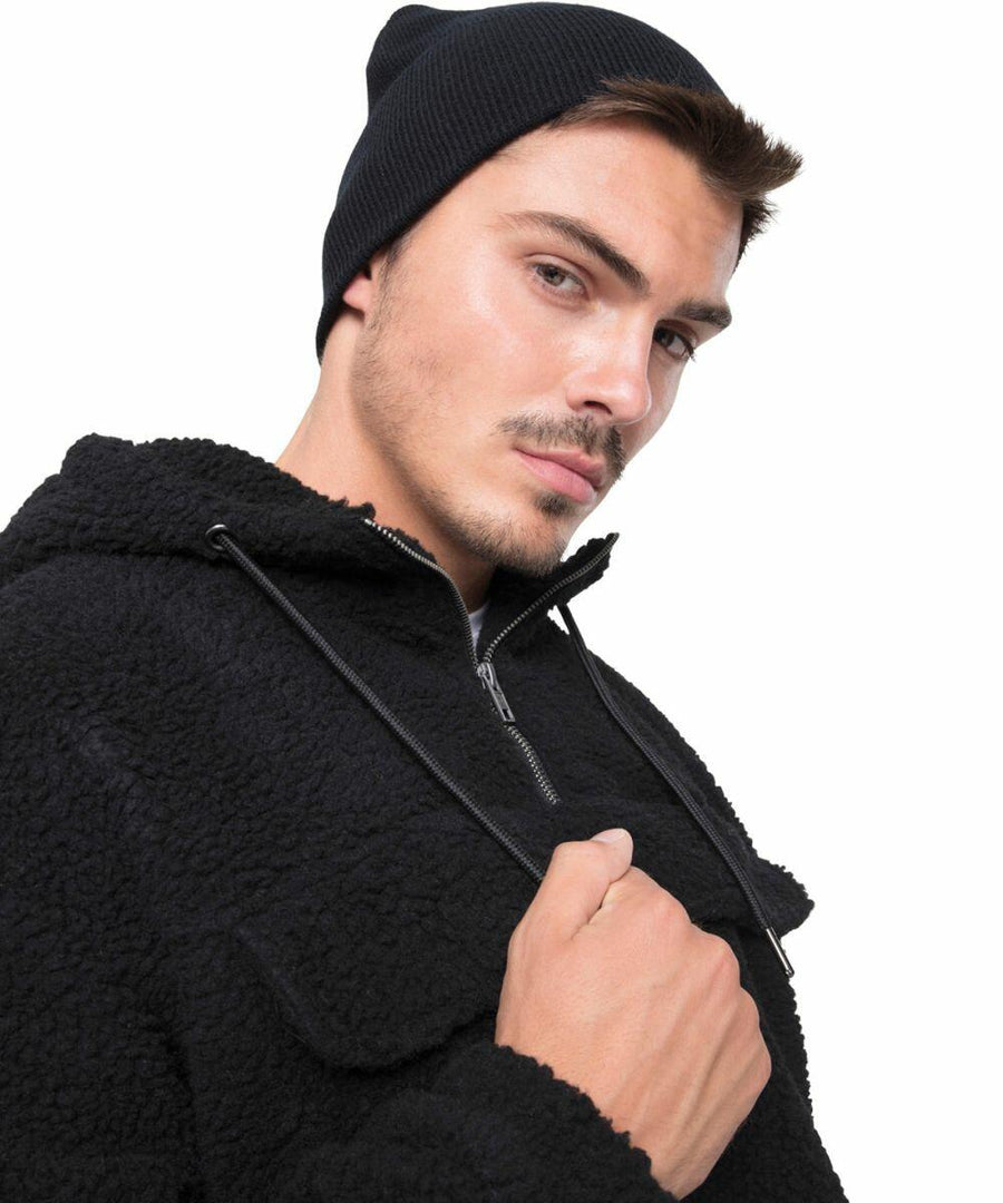 A man wearing a black Flexfit By Yupoong YP013 beanie and a black hooded jacket stands against a plain white background.
