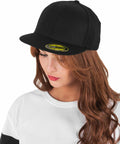 A person with long brown hair wearing a black Flexfit By Yupoong YP017 baseball cap looks downwards. They are dressed in a white and black top.