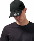 Person wearing a black T-shirt and a black Flexfit By Yupoong YP022 baseball cap with a sticker on the brim. The person is looking downward.