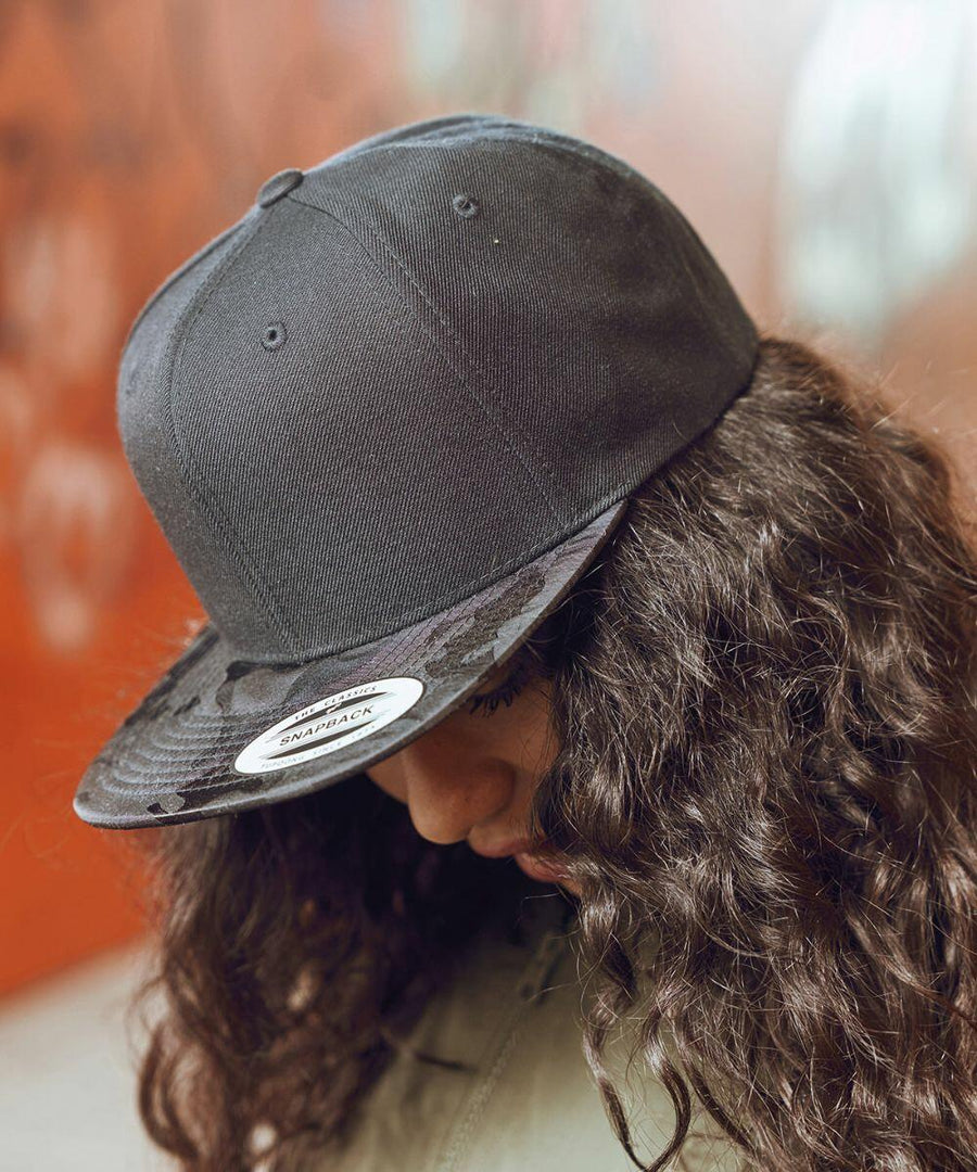 Person with long curly hair wearing a black Flexfit By Yupoong YP025 cap looking downward.