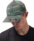 A person wearing a green camouflage **Flexfit By Yupoong YP026** cap and a black shirt looks downwards.