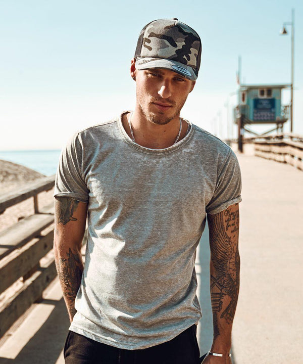 Man wearing a gray t-shirt and Flexfit By Yupoong YP029 camouflage baseball cap stands on a pier with a lifeguard tower in the background on a sunny day.