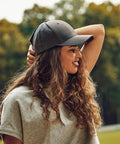 A woman with long, curly hair wearing a gray Flexfit By Yupoong YP035 baseball cap and gray shirt stands outdoors, looking to the side, with one hand on her cap. Trees are visible in the background.